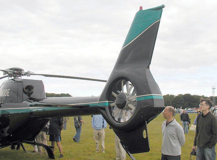 Photograph of a Fenestron shrouded tail rotor on a light helicopter: multiple short blades set inside a duct through the vertical fin, with bystanders beside it for scale.