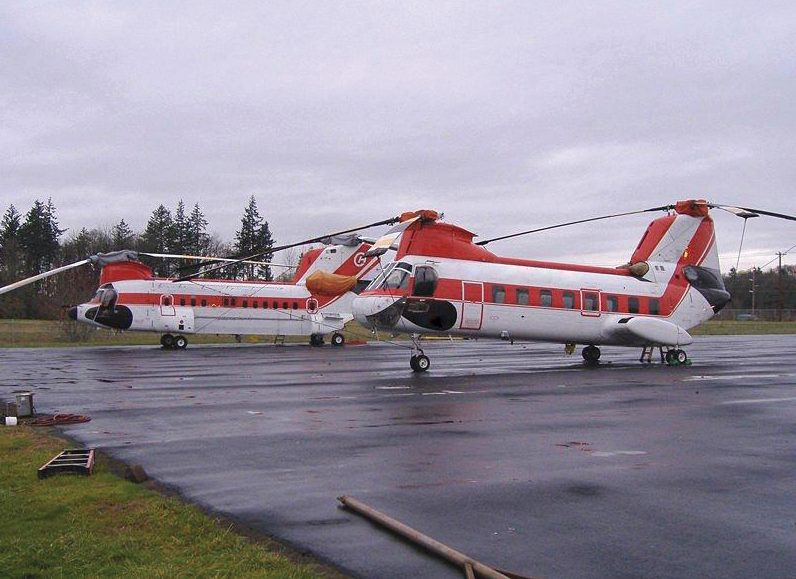Two tandem-rotor helicopters parked on a wet ramp, each with a large rotor at the front and another at the rear and no tail rotor.